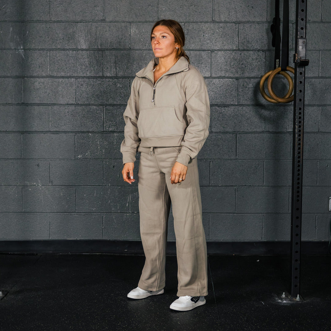 A woman standing in a casual pose wearing gray sweatpants and a matching hooded sweatshirt, with white sneakers and a subtle embroidered logo on the pants.