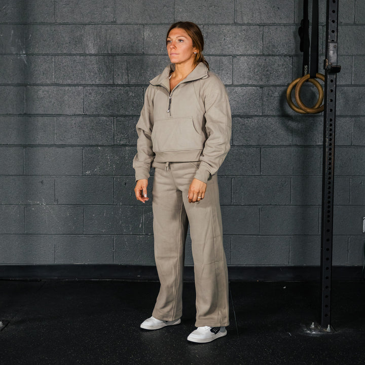 A woman standing in a casual pose wearing gray sweatpants and a matching hooded sweatshirt, with white sneakers and a subtle embroidered logo on the pants.