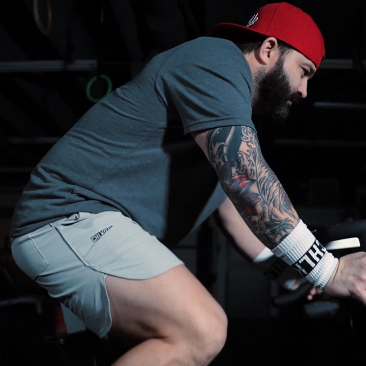 Man riding an assault bike in a dark gym, wearing white FLX Pro Shorts and a red cap.