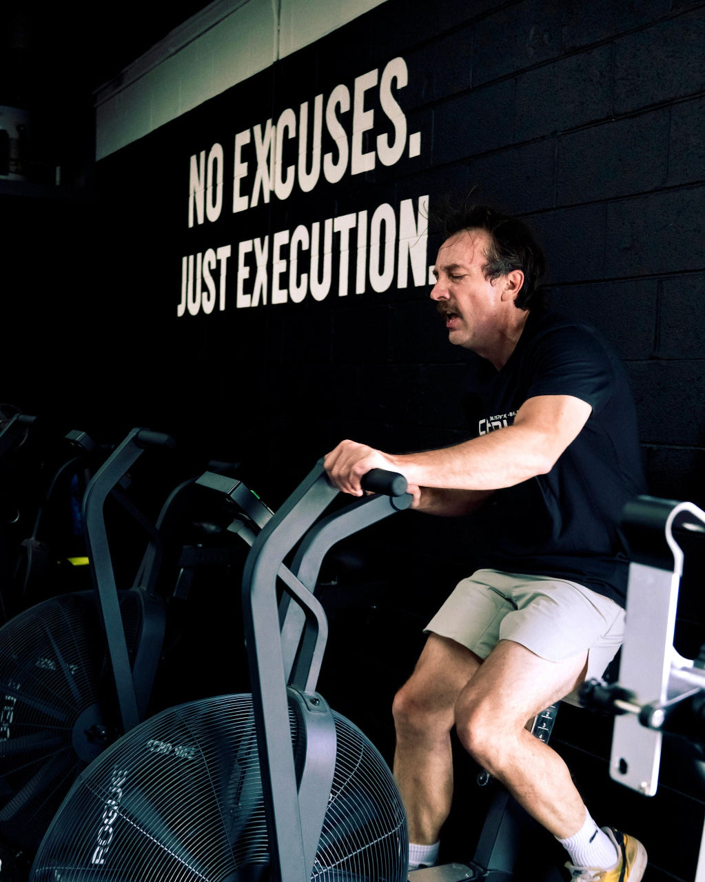 Man exercising on a stationary bike with 'No Excuses. Just Execution.' motivational quote on the wall.